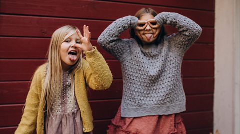 Two girls pulling silly faces. One blonde girl wearing a pink dress and yellow cardigan sticks her tongue out and gestures with her hand to her nose. Another brunette girl next to her in a pink skirt and grey jumper sticks her tongue out for the camera and puts her hands to her face to make it look like she is wearing glasses