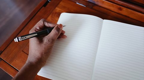 A left hand holds a black pen as it is about to start writing in a white lined notebook on a wooden table