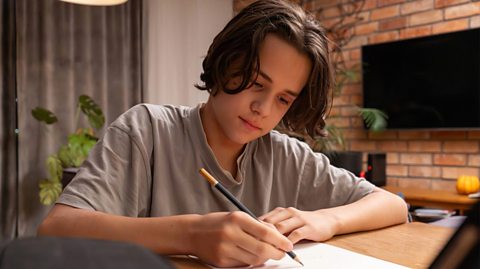 A teenage boy with longer brown hair and wearing a grey t-shirt sits at a wooden table writing on paper. He holds a dark blue pencil in his right hand.