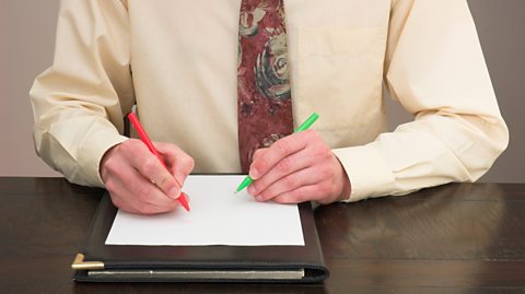 A man wearing a yellow shirt and dark red patterned tie sits at a wooden desk. He holds a red pen in his right hand and a green pen in his left as he attempts to write with both hands on a piece of paper