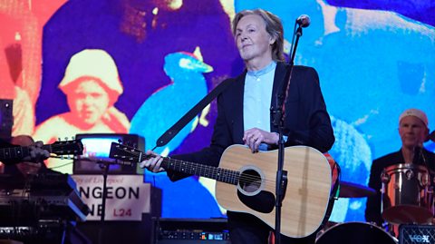 Legendary artist Sir Paul McCartney has longer grey hair and wears a blue shirt and navy blue blazer on stage with his guitar