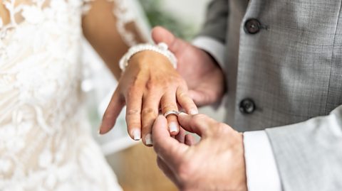 A woman in a white lace wedding dress holds out her left hand as her husband, who wears a grey suit, places a ring on her wedding finger