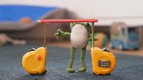 A toy frog stares at a pencil supported by two tape measures, ready to limbo beneath it.