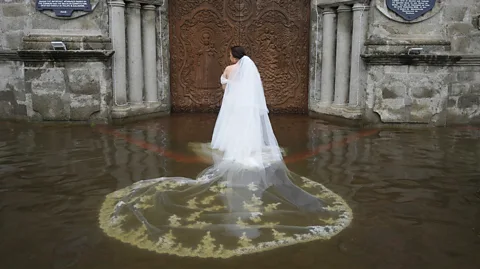 Aaron Favila/ Associated Press A woman wearing a wedding dress and a soaked veil stands, with her back to the camera, in brown floodwater in front of a large wooden church door in the Philippines (Credit: Aaron Favila/ Associated Press)