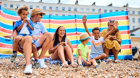 mixed race family sit on a pebbly beach in front of a windbreak enjoying the summer