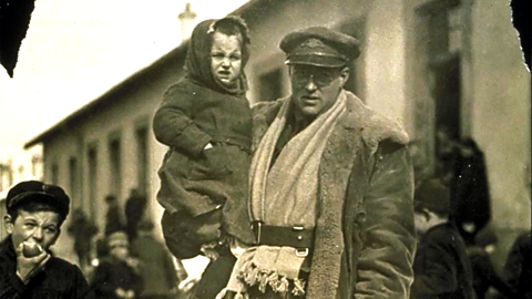 First World War airman in uniform carrying a child along a street, with other people visible in the background.