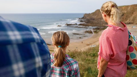 Parents and their daughter look at a cove from a clifftop - seeing if the beach is safe to swim.