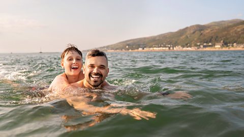 Dad and son in the sea off Wales engage in some fun splashing around in swimwear.