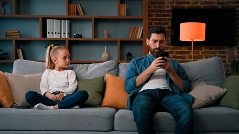 annoyed child eyes father as he is engrossed in his smartphone on a sofa