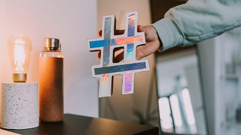 A hand holds out a large silver and multicoloured hashtag symbol - a small square grid made of four intersecting lines: two straight horizontal lines stacked on top of each other, crossed by two vertical. There is also a desk, bulb lamp and a flask