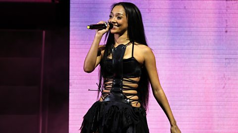 KATSEYE's Manon smiling while performing on stage. She is wearing a black dress and holding a black microphone. 