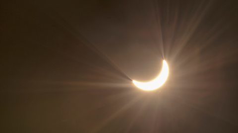 A landscape image shows the night sky, which is cast in a purple-ish hue. In the centre, the sun forms a crescent shape because the moon is partly covering it in a partial eclipse. 