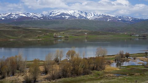 a landscape image shows a lake surrounded by snow-capped mountains. Trees and meadows dot the shore under a blue sky with scattered clouds.