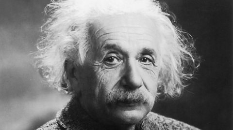 A black and white, head and shoulders photograph of Albert Einstein. He's an older white man with a bushy moustache and grey hair that looks slightly dishevelled and unkempt. He's looking directly at the camera. 