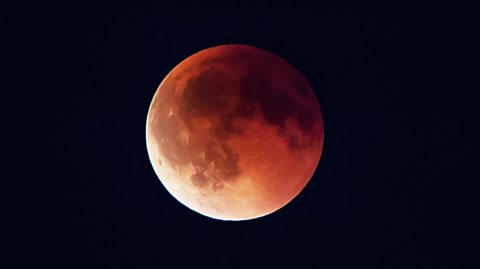 The image shows the pitch black night sky. In the centre is the moon, cast in an eerie red light during a lunar eclipse. 
