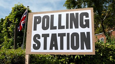 The image shows a large wooden sign with bold, block-capital black lettering that reads 'polling station'. There are trees in the background against a blue sky. 