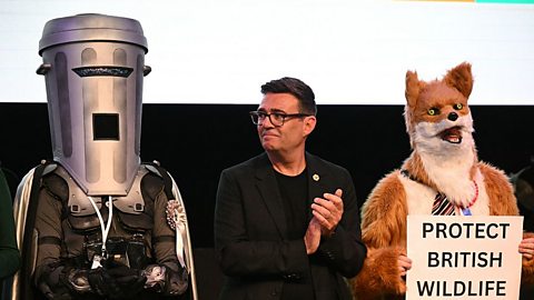 An image of Count Binface, Andy Burnham and a candidate dressed in a fox costume holding a sign that reads 'protect British wildlife', at the Makerfield by-election count in 2026. 
