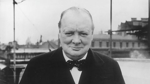 A black and white photograph of Winston Churchill. He's looking directly at the camera and giving a closed-lipped smile, wearing a black suit with a black bowtie. 