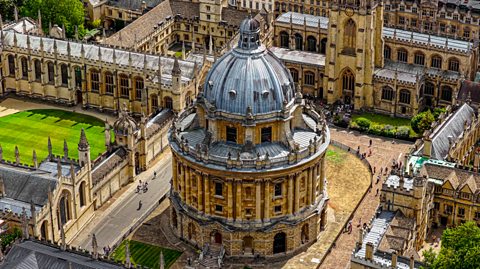 A view of Oxford from above. In the centre is the Radcliffe Camera, an impressive circular building with a domed roof. Around it are other university buildings with courtyards, spires and manicured lawns. 