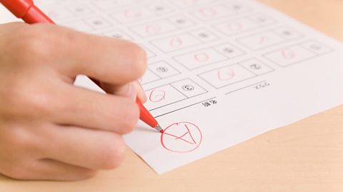 A close-up image of an upside-down exam paper on a table. A hand holding a red pen is writing the letter 'A' in the top corner and circling it. 
