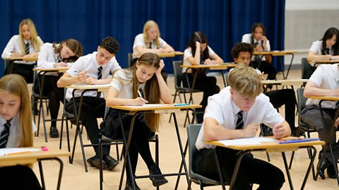 Rows of students sitting at narrow fold-out desks are taking exams in a school hall. 