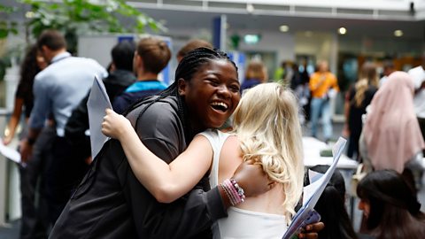 The image shows two female friends hugging each other while holding their exam results slips. There are lots of people milling around in the background. The girl whose face we can see is laughing and appears very happy.