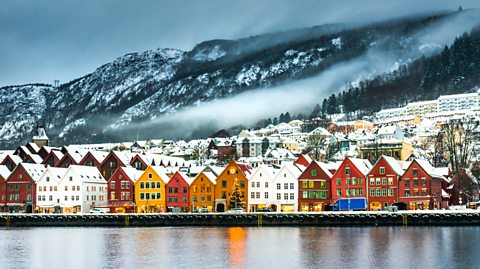 A wintery scene in Bergen, Norway, showing coloured houses with snowy rooftops lining the waterfront, with mountains in the background