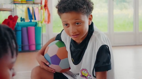 A boy with a football under his right arm talks to another child in a classroom