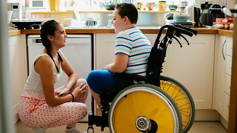Older sister chats to younger brother who is a wheelchair user as they sit in a sunny kitchen