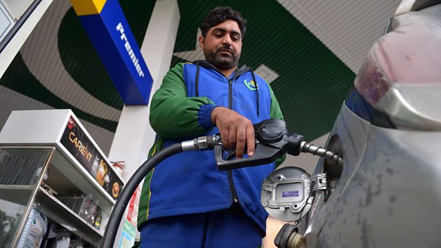 Man refuelling a car at a petrol station, holding a fuel nozzle connected to the vehicle, with pumps and station signage visible in the background.