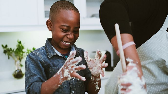 A child looks at his hands which are covered in dough and he's smiling