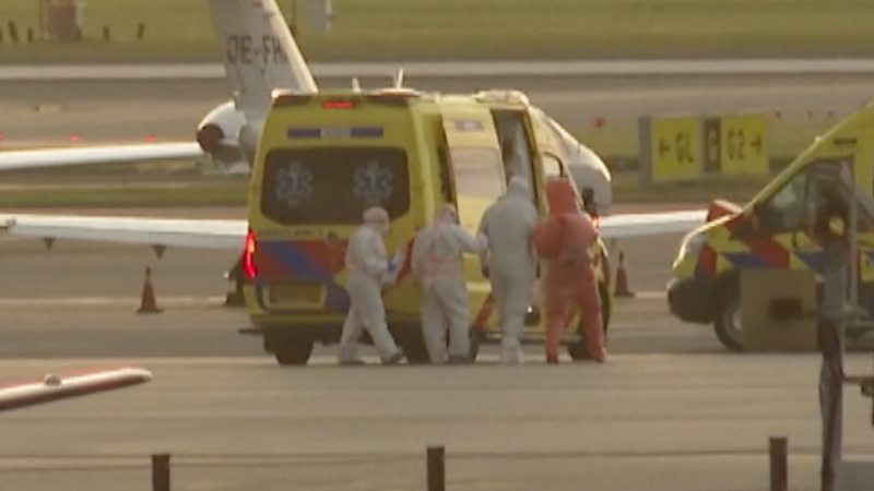 People in white and orange hazmat suits walk to a yellow ambulance at an airport