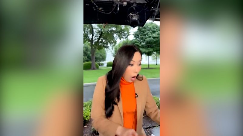 A reporter dressed in an orange turtleneck jumper and beige blazer looks shocked and gasps as she starts to run.
