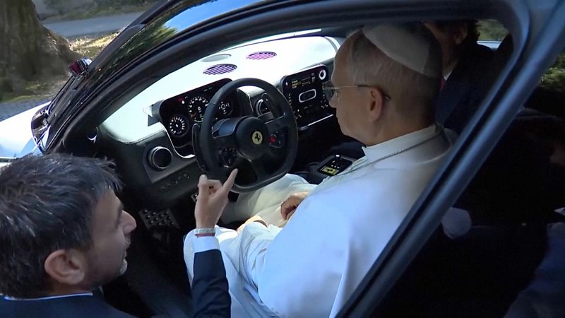 The pope dressed in a traditional white Cassock sits in a Ferrari with another man pointing to the steering wheel.