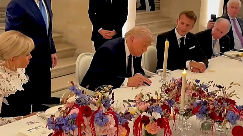 Trump signs a paper next to Macron and others on a decorated table