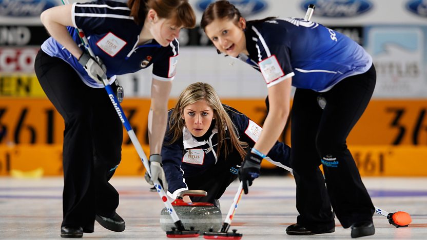 Scottish curler Eve Muirhead crouches in a lunge position in the ice, watching as two sweepers brush the ice in front of her red stone. All are wearing navy.