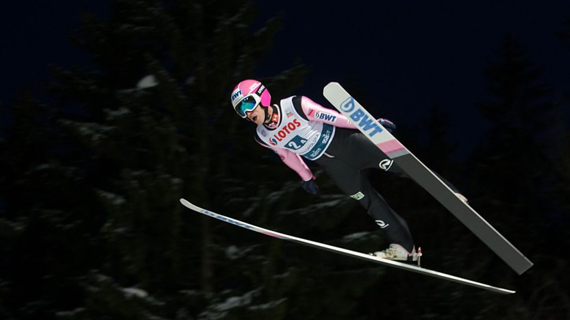 A ski jumper wearing a pink helmet and jumpsuit soars through the air, with his skis held in a V position beneath him