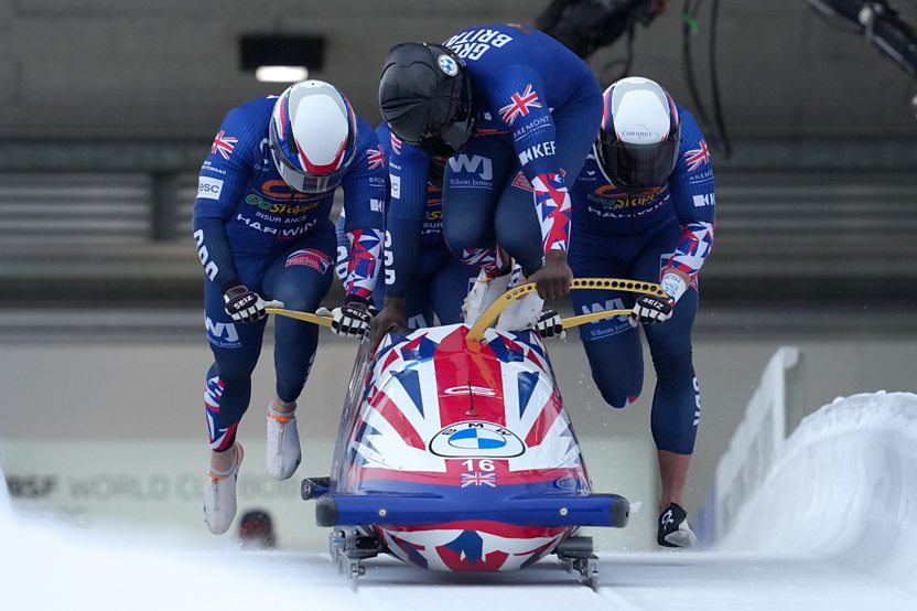 Four athletes wearing tight-fitting navy jumpsuits push a bobsleigh along an ice track. The first athlete is halfway through jumping into the sled.