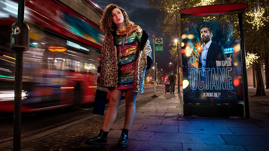 Rose Matafeo walks past a poster showing Nikesh Patel in a fictional Hollywood film