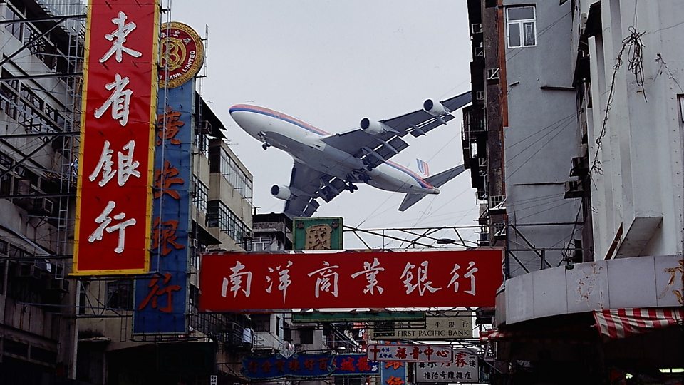 A plane going in for landing low over a Hong Kong street full of signs.