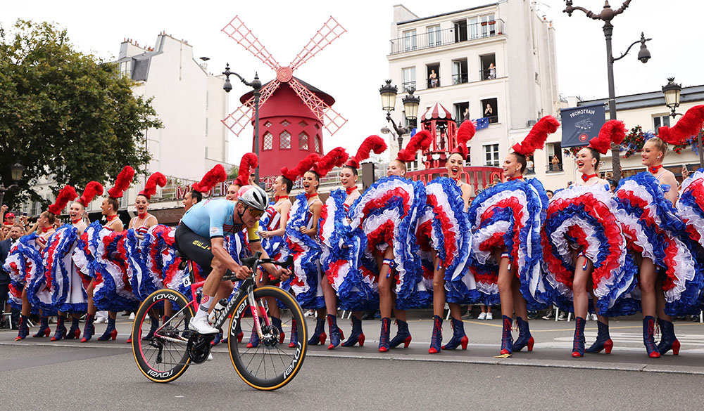 Remco Evenepoel from Belgium, cycles through the Moulin Rouge with Can Can dancers performing in the background