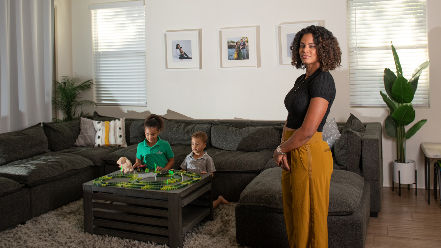 Allison Mc Cullough stands in her living room while her children, a daughter and a son, play a game sitting at a table on the background.