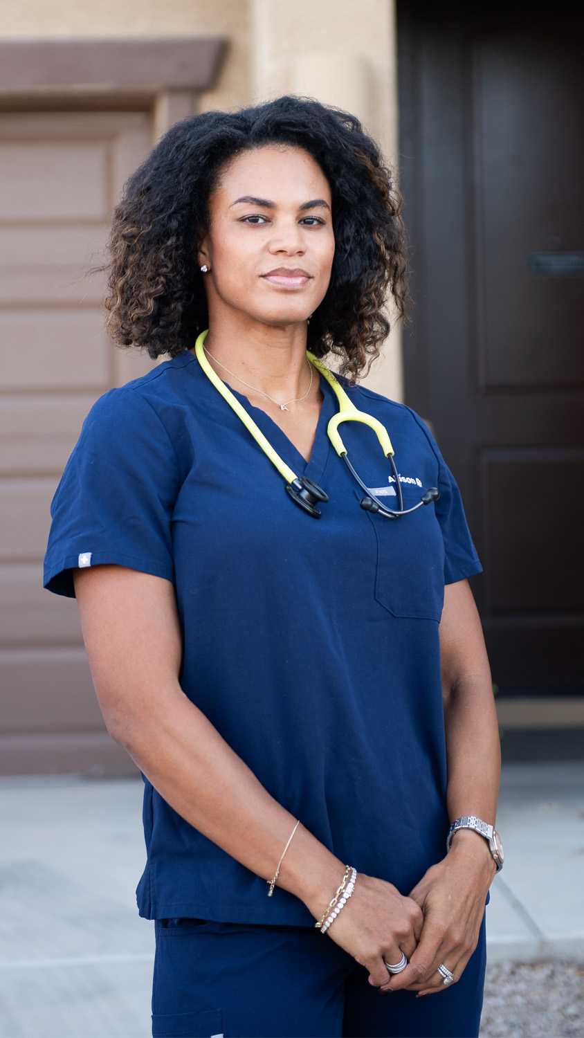 Allison Mc Cullough stands in front of her house dressed in a doctor's uniform, blue scrubs.