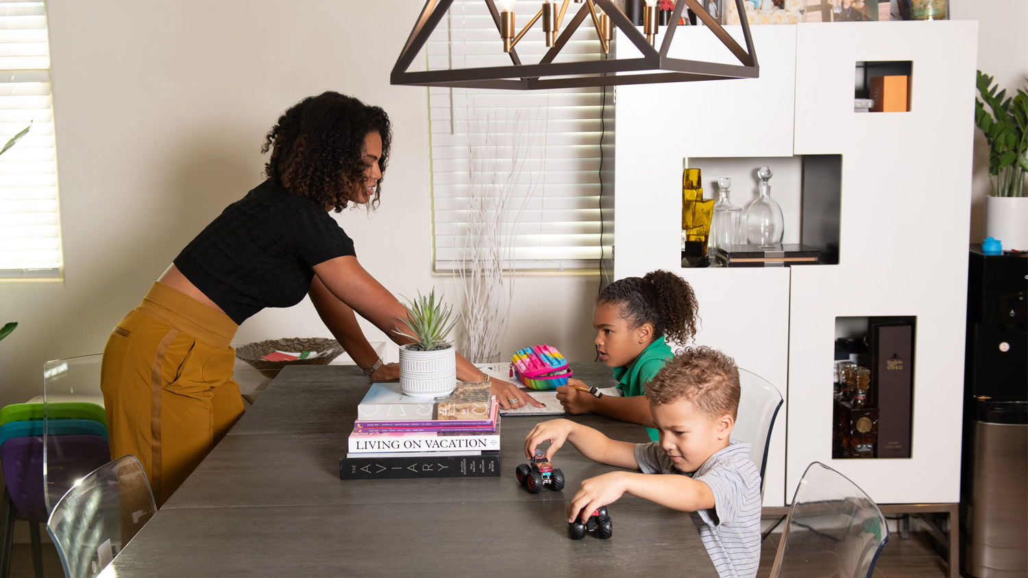 Allison McCullough plays stands over a kitchen table and shows something to her daughter in a book while her son plays with toy cars.