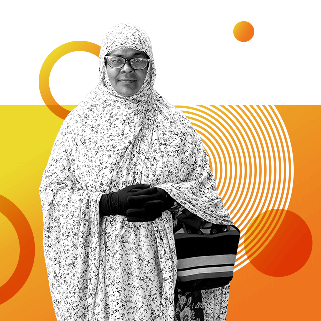 Nurse and school founder Rikta Akter Banu in a full length floral chador and headscarf, wearing gloves and clutching her hands together in front of her, with a stripy handbag around her wrist.