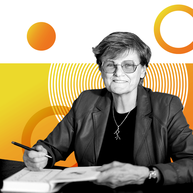 Katalin Karikó, biochemist and Nobel laureate, with glasses on and smiling to camera, sitting behind a desk signing a book, with short hair, wearing a jacket and a necklace.