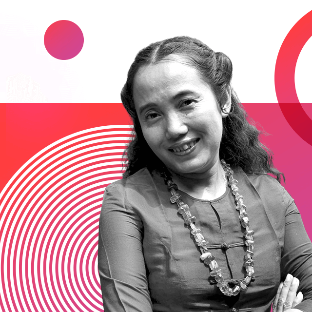 Film-maker Shin Daewe, smiling, wearing her long hair tied half back and a Myanmar-style cotton top with buttons straight down from the neckline and a chunky beaded necklace whilst crossing her arms.