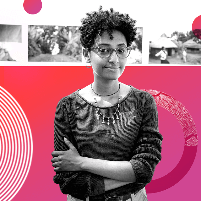 Photographer Maheder Haileselassie wears a loose fitting knitted jumper with three stars on it, assorted necklaces and clear rimmed glasses. She stands with her arms folded around her torso and she looks at the camera, while three of her photos hang in the background, out of focus.