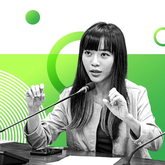 Politician Huang Jie, with a fringe, sitting behind a desk talking into a small microphone, as if presenting to an audience.