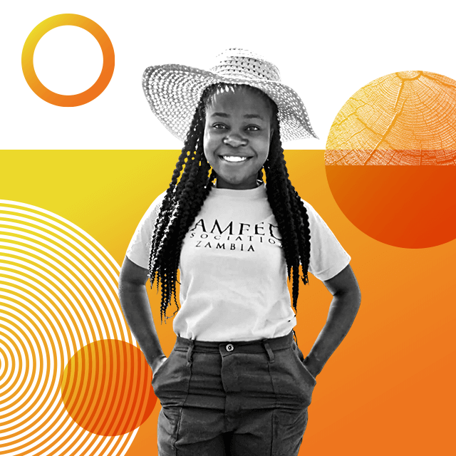 Farmer Naomi Chanda wearing a straw hat and a T-shirt that says “Camfed Association Zambia”. She has braided long hair and is posing with her hands in her trouser pockets.